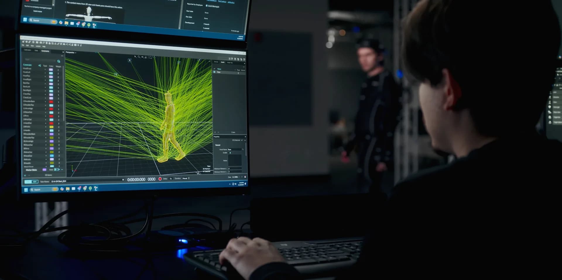 Person at a monitor running OptiTrack motion-capture software: a tracked skeleton with camera rays, a person in a marker suit in the capture volume behind