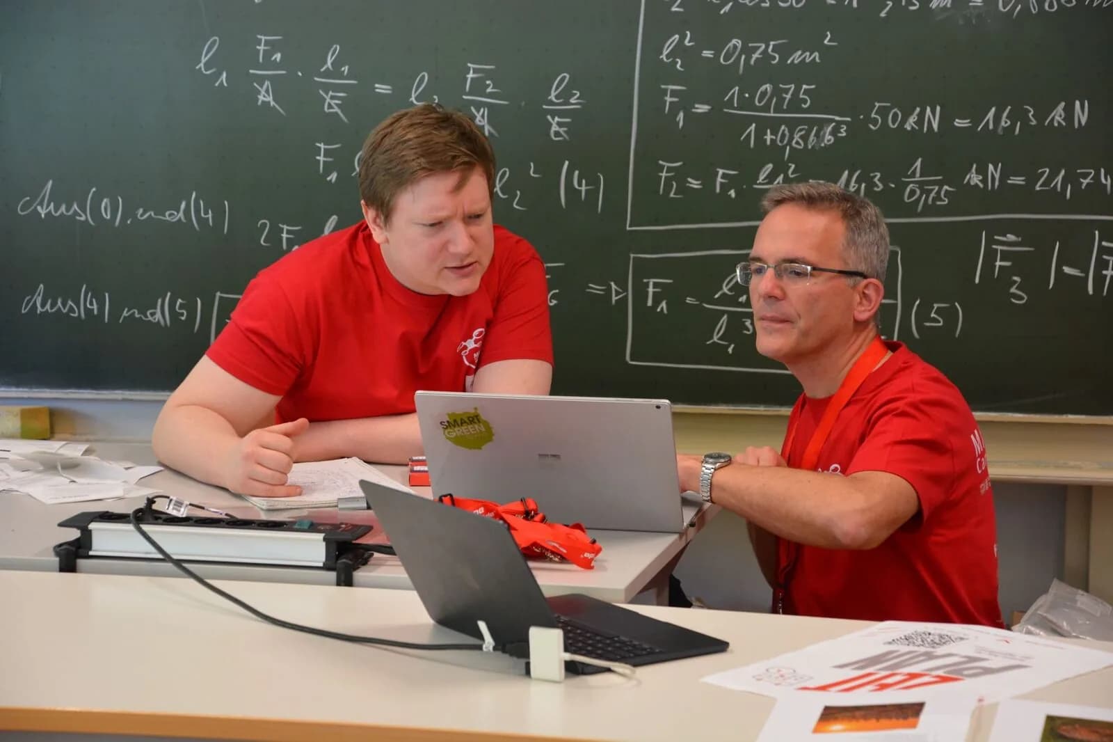 Zwei Teilnehmer tüfteln gemeinsam am Laptop vor einer Tafel mit Berechnungen